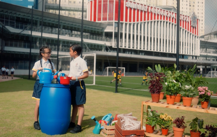 Two Sunway International School children at the Sunway International School football field, alongside plants and a water tank, with kids playing football in the background